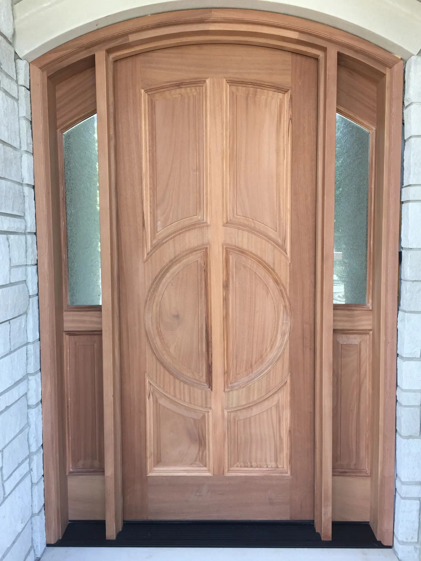 An arched entry door in bare wood with glass sidelights, before staining, set in a stone surround.