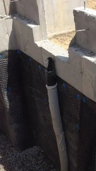 A foundation wall faced with black dimpled membrane and a drain pipe running down it, with gravel at the base.
