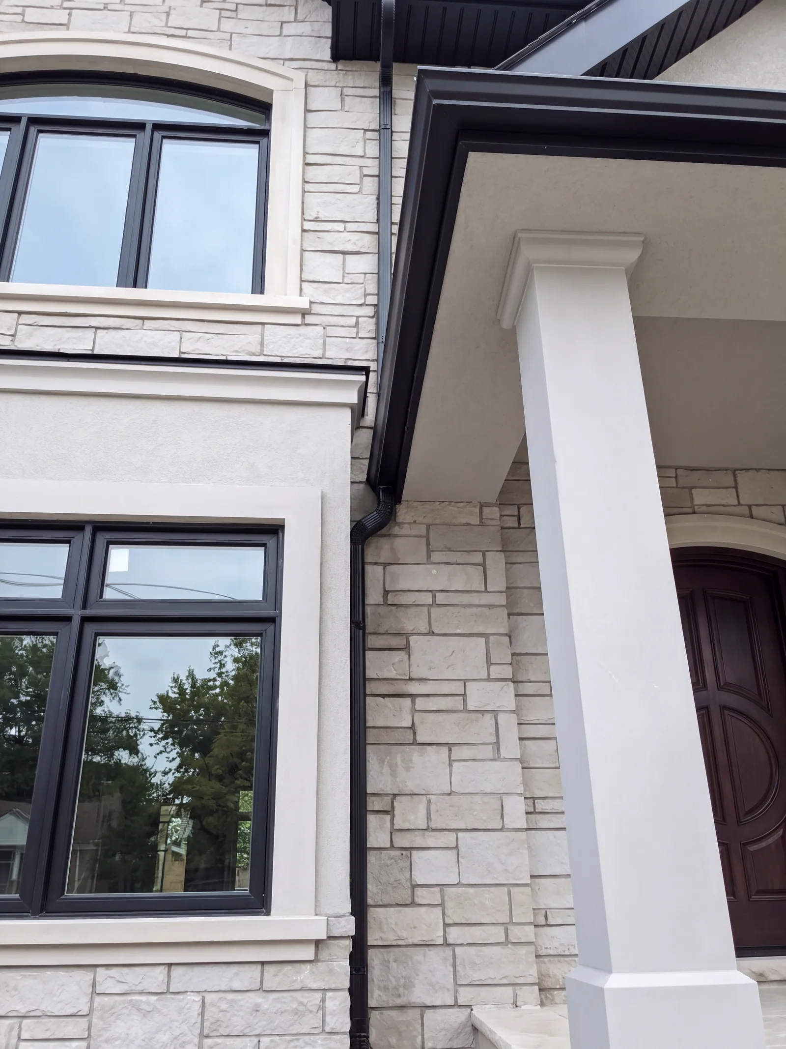 A square white porch column and the roof edge of a stone-clad house, beside a dark-framed window and the edge of the arched front door.