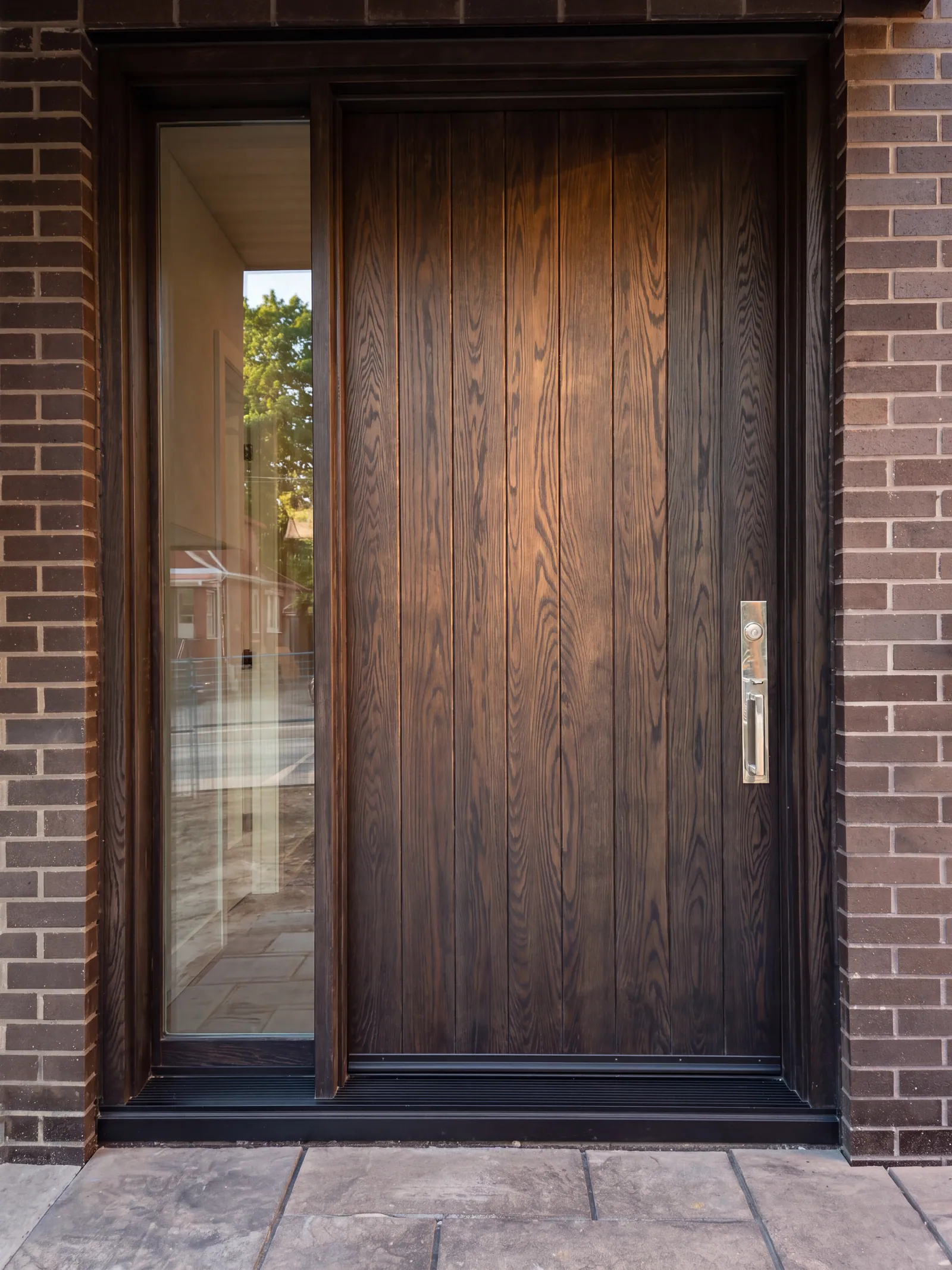 A tall plank front door in dark stained wood with a tall glass sidelight, in a brick surround, above a stamped concrete landing.