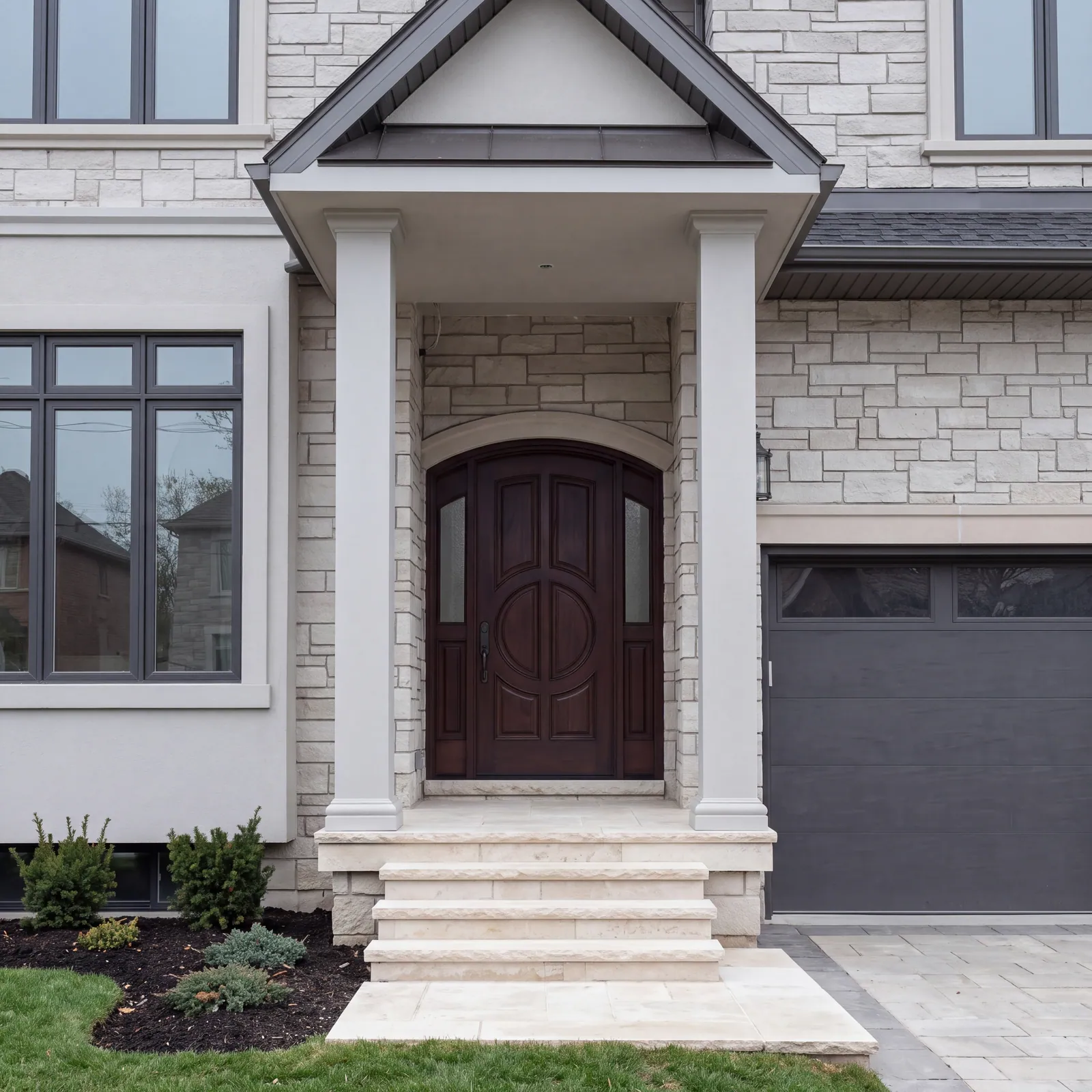 The front porch of a stone house, with a gabled roof on two square white columns, an arched dark wood door with sidelights, stone steps and a dark garage door beside it.
