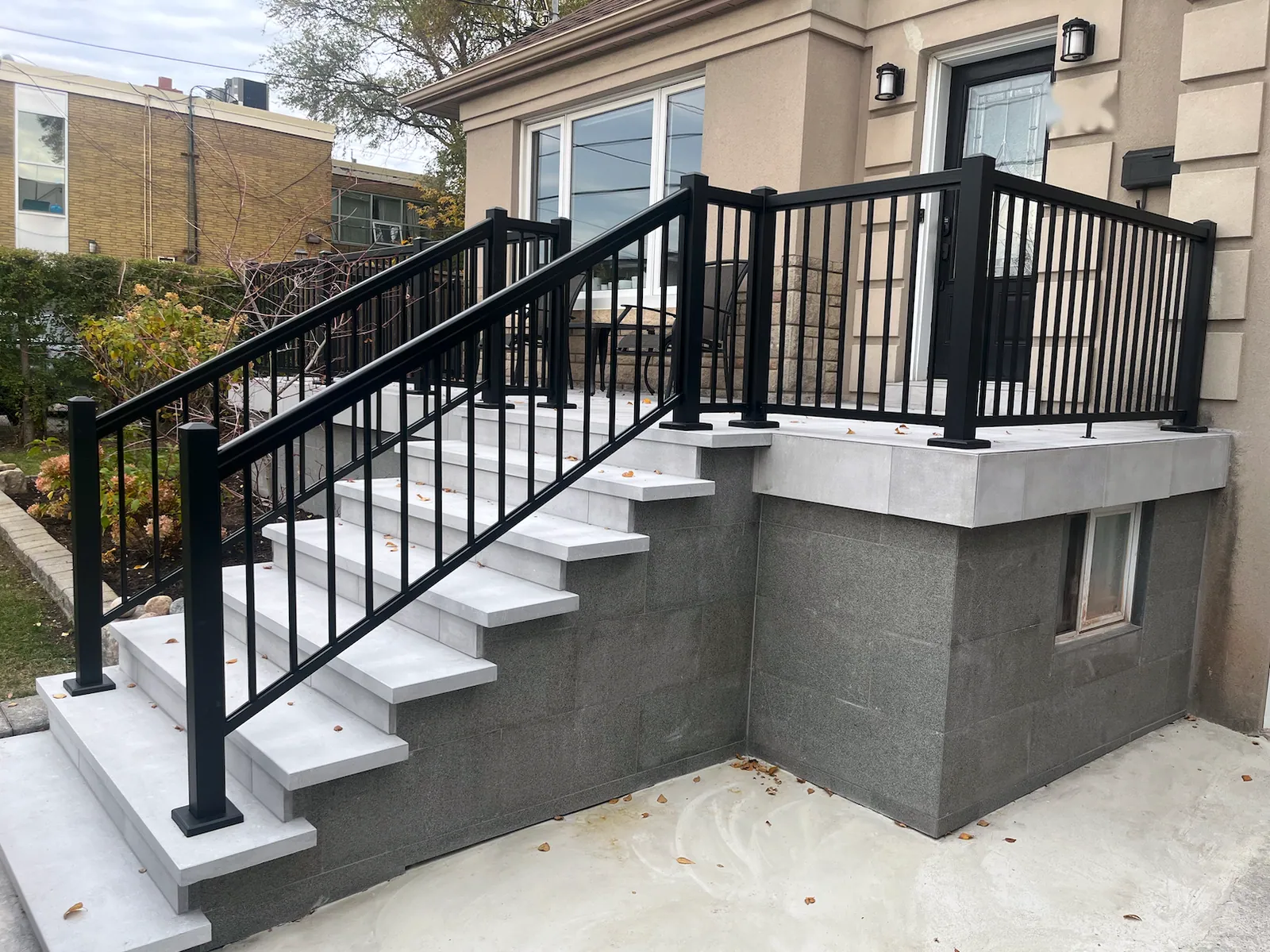 Pale grey stone steps with a black metal railing rising to a landing with a black metal guard, in front of a stucco house with a grey block base and a small basement window.
