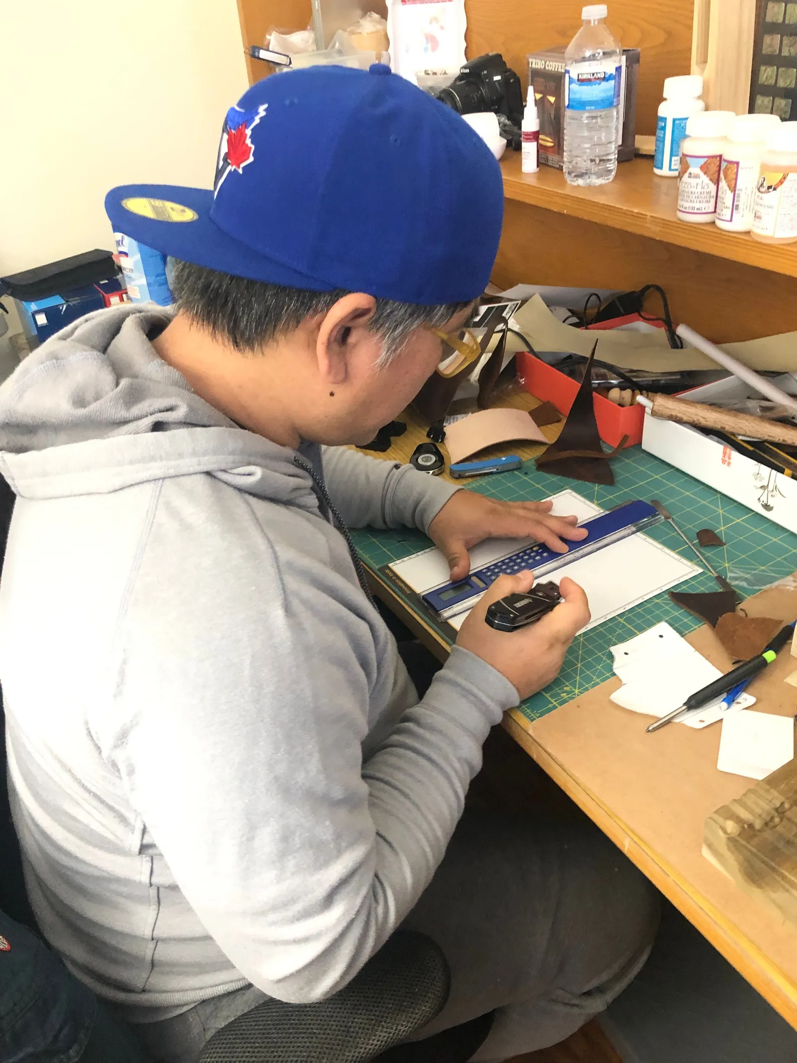 Young in a blue cap at a desk, cutting and stitching leather.