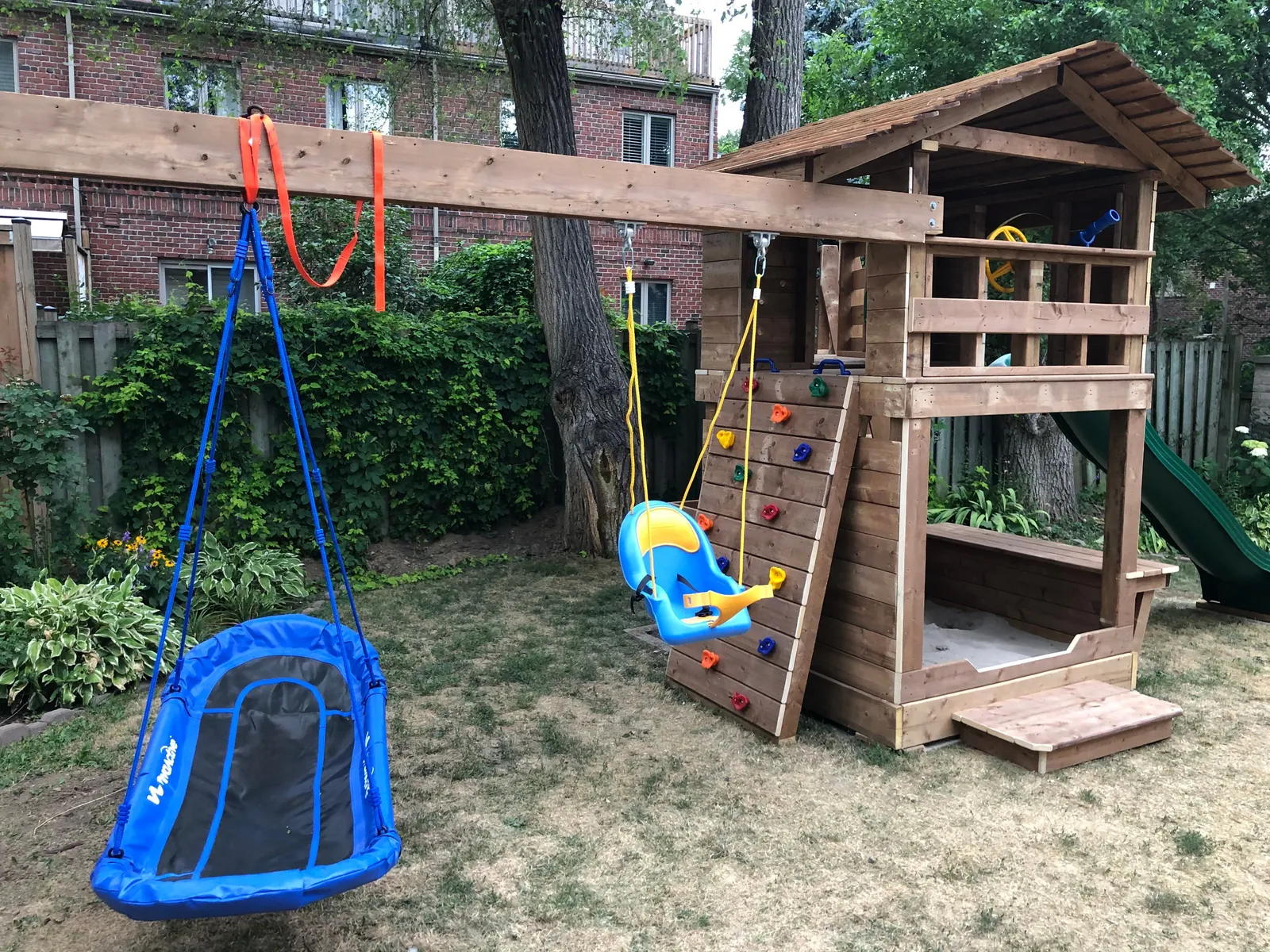 A wooden play fort with a green slide, a swing beam and a blue nest swing.