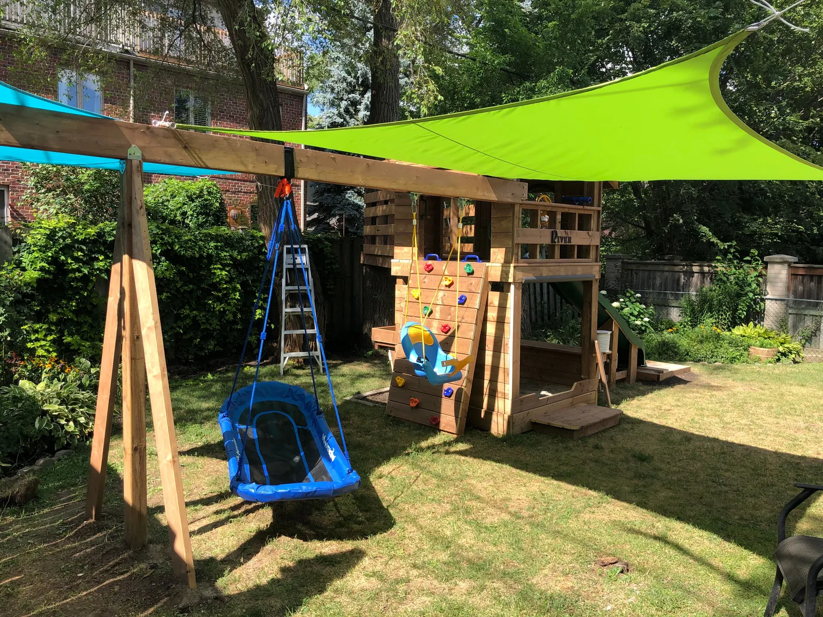 The play fort with a green shade sail, A-frame swing posts and blue swings.