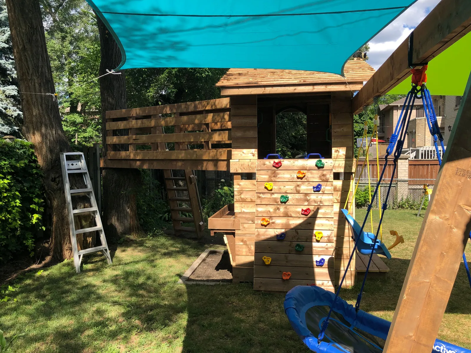 The play fort with a teal shade sail, a ladder, a climbing wall and a stone base.
