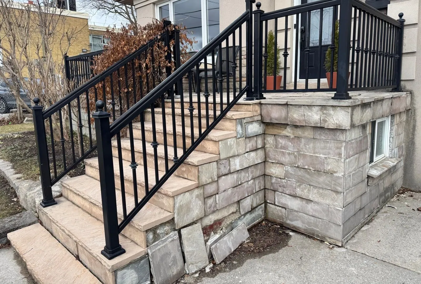 Stone steps with a black metal railing at the front of a house, beside a stone-faced wall with a small basement window.