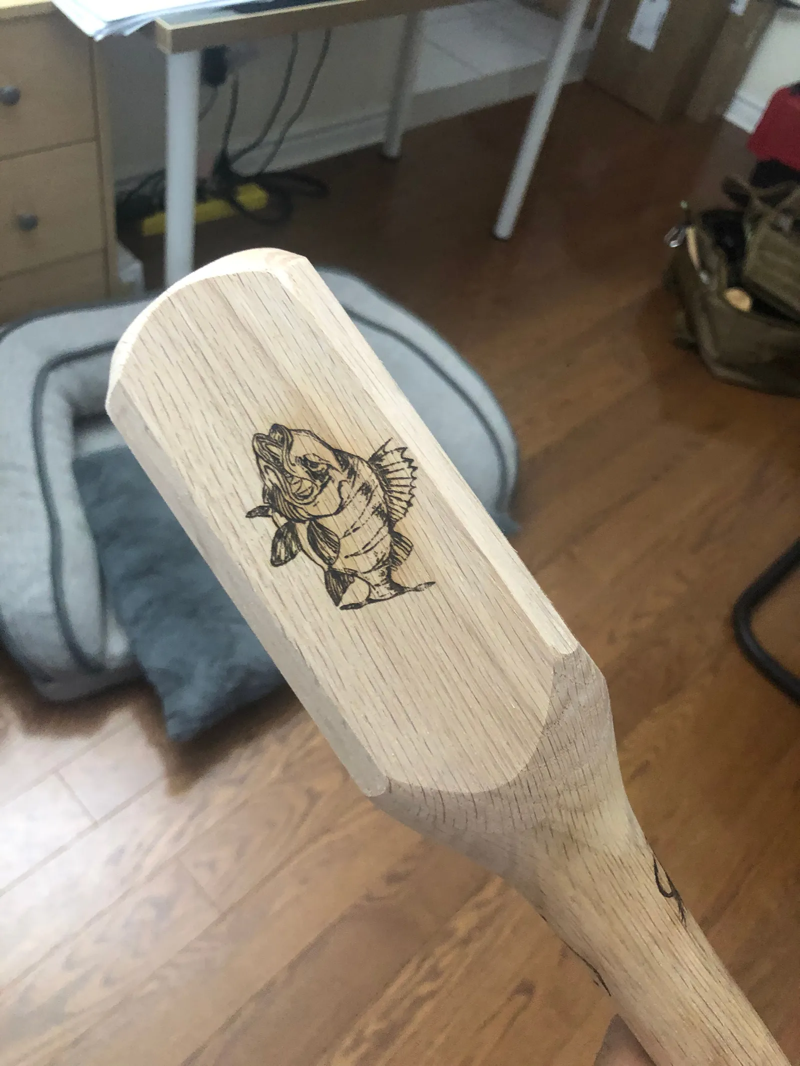 A wooden paddle-shaped piece engraved with a fish, lying on a floor.